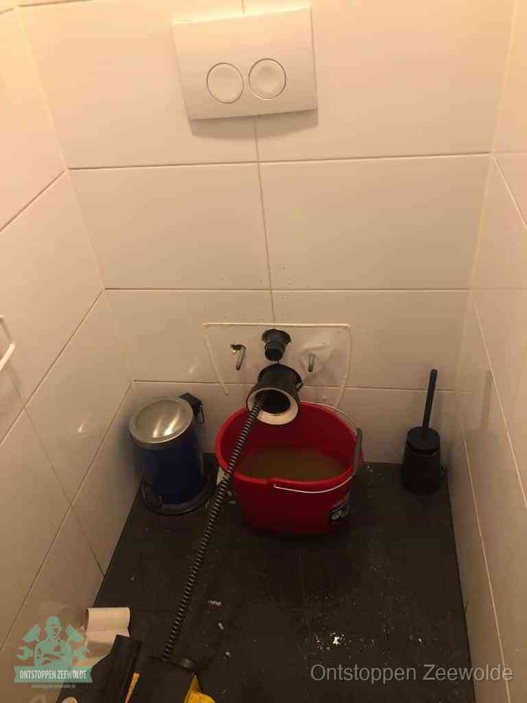 Een ontstopte wand-wc met verwijderde toiletpot waar een ontstoppingsveer in de wanduitgang verdwijnt, het achterhalen waar de verstopping zit in Zeewolde