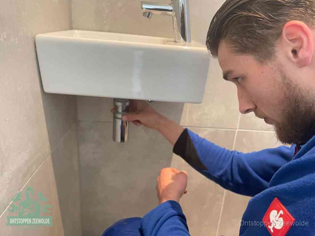 Een monteur draait met de hand de zwanenhalssifon onder een fonteinwasbak in de toiletruimte los om de bron van rioollucht in Zeewolde te vinden