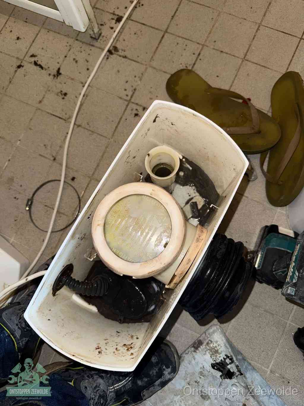 Een gedemonteerde toiletpot op de vloer met ontstoppingsveer en gereedschap ernaast, het verwijderen van een vastzittend voorwerp uit de wc in Zeewolde