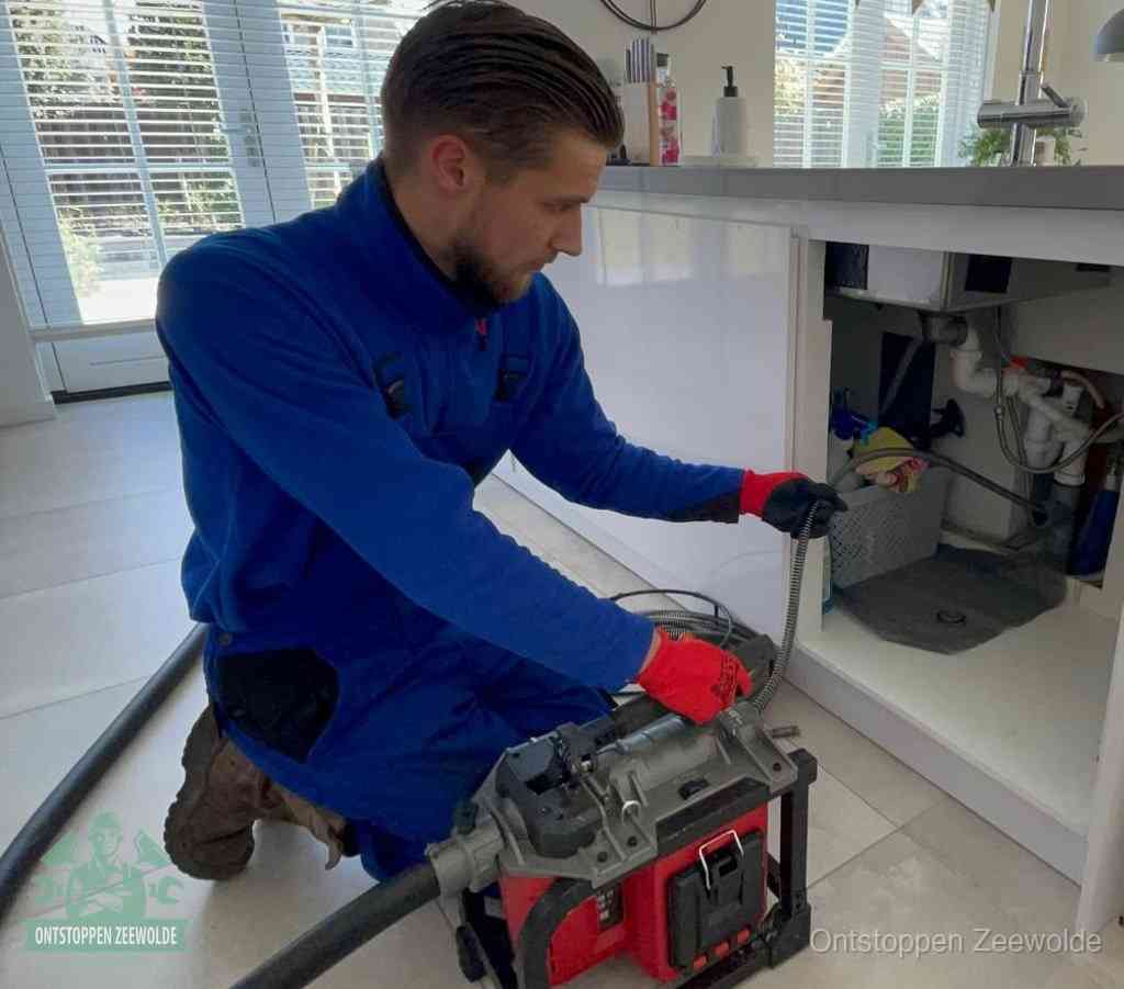 Een monteur met rode handschoenen knielt in een keuken en voert een elektrische ontstoppingsveer in tijdens het professioneel afvoer ontstoppen in Zeewolde